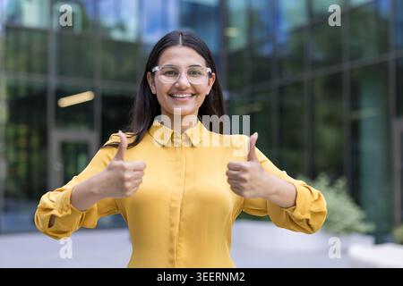 Junge Geschäftsfrau mit Brille und gelbem Hemd, steht draußen und zeigt selbstbewusst zwei Daumen mit einem fröhlichen Ausdruck, symbolisiert Anerkennung und Erfolg Stockfoto