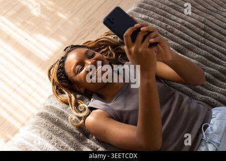 Afroamerikanerin im grauen Tank, kurze Hosen, auf grauem Überwurf auf dem Bett liegend und lächelnd am Telefon Stockfoto