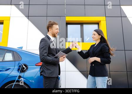 Glückliche Kundin, die vor einem brandneuen Auto steht und die Schlüssel vom Händler im Außenbereich des Händlers abnimmt. Die Frau ist sehr zufrieden mit dem neuen Auto, das sie hält Stockfoto