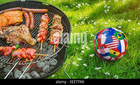 Köstliches gegrilltes Fleisch mit Gemüse, das auf dem Grill über den Kohlen brutzelt, mit einem Fußball bedeckt mit mehreren Nationalflaggen, die einen internati symbolisieren Stockfoto