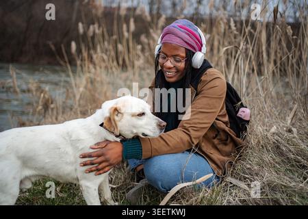 Glückliche junge Erwachsene Frau, die in der Natur Hund streichelt und Musik hört Stockfoto
