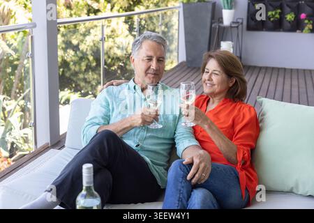Seniorenpaar, das auf einem Balkonsofa sitzt und mit Weingläsern in der Nähe einer Weinflasche getoast hat Stockfoto