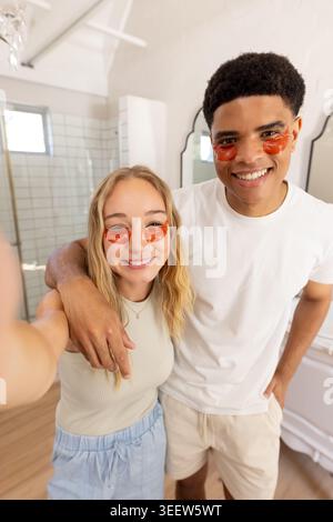 Ein Paar lächelt und nimmt Selfie im Badezimmer, trägt Gel-Patches unter den Augen und legere Kleidung Stockfoto