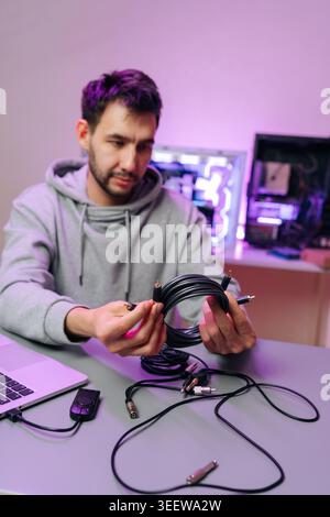 Mann in Kapuzenpullover, der konzentriert Audio- und Datenkabel an einem unordentlichen Schreibtisch sortiert und inspiziert, während er Verbindungen zwischen Laptop und benutzerdefiniertem RGB-beleuchtetem PC vorbereitet Stockfoto