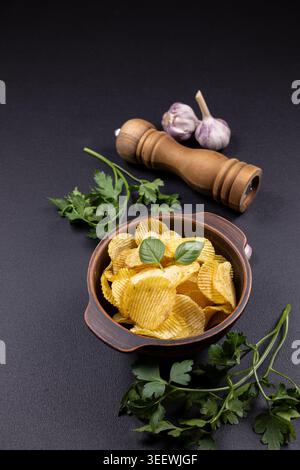 Appetitliche Kartoffelchips, die in einer rustikalen braunen Keramikschale serviert werden, garniert mit frischen Basilikumblättern, Knoblauchzwiebeln, Petersilie und einem Holz Stockfoto