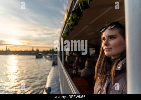 Kaukasische Frau, Die Über Den Sonnenuntergang Am Fluss Blickt, Sich Auf Das Geländer Des Waterfront Cafe Bootes Lehnt, Warmes Goldenes Licht, City Skyline Im Hintergrund, Sonnenbrille Stockfoto