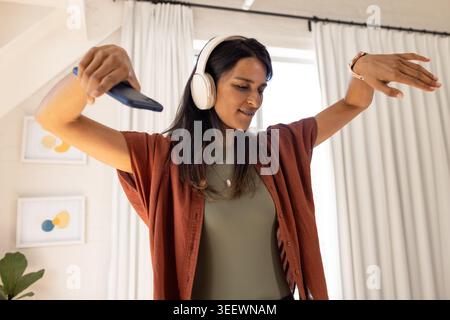 Indische Frau tanzt und hört Musik mit Kopfhörern, während sie das Smartphone im Loft hält Stockfoto