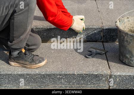 Arbeiter Kniet Auf Betonstufen Und Repariert Beschädigte Gelenke Mit Handschuhen, Wobei Kelle Und Meißel In Der Nähe Von Schaufel Und Rauer Asphaltkante Verwendet Werden, Fokussiert Stockfoto