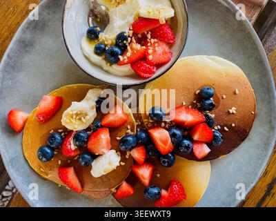 Hausgemachte Pfannkuchen mit frischen Erdbeeren, Heidelbeeren und Nüssen, serviert mit cremigem Joghurt. Rustikaler Holzrücken Stockfoto