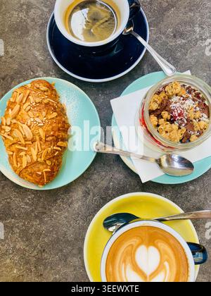 Hochwinkel-Aufnahme eines köstlichen Frühstücks: Cappuccino mit herzhafter Latte Art, schwarzer Kaffee, Mandelcroissant und Müslijoghurtschale Stockfoto