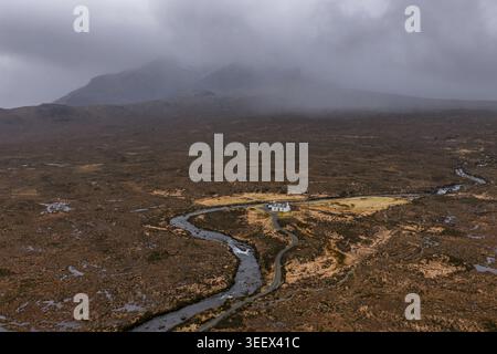 Aus der Vogelperspektive auf ein einsames weißes Cottage eingebettet von einem gewundenen Fluss in der weiten Moorlandschaft unter nebeligen Bergen Isle of Skye, Schottland, United K Stockfoto