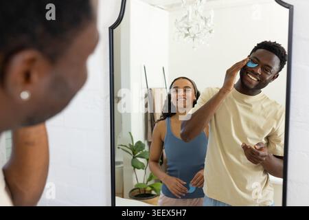 Afroamerikanisches Paar, das blaue Gesichtspads aufträgt und den Kosmetikspiegel im Badezimmer mit einem Gläser für Hautpflegeprodukte benutzt Stockfoto