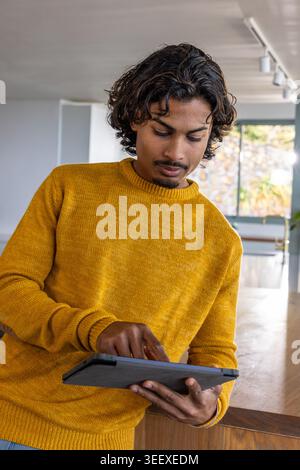 Asiatischer Mann, der an der Holztheke in der Küche steht, trägt Senfpullover, der Tablette im Koffer klopft Stockfoto