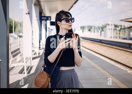 Die junge Frau hält das Smartphone, während sie auf dem sonnigen Bahnsteig steht. Moderne Bahnverbindungen, öffentliches Verkehrsnetz, nachhaltiges Stadtverkehr Stockfoto