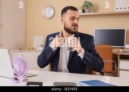 Junger Geschäftsmann mit Glas Wasser und Mini-Ventilator, der am Tisch im Büro unter Hitze leidet Stockfoto