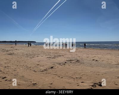 Riga, Lettland - 2. Mai 2026: Weite Küstenlandschaft, in der Menschen entlang eines Sandstrandes unter einem hellblauen Himmel mit Kondensstreifen über dem Meer spazieren Stockfoto