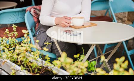 Frau sitzt im Café im Freien und hält eine Tasse Kaffee, Smartphone auf dem Tisch, gemütliche urbane Umgebung mit Stühlen und Pflanzen, Konzept der Entspannung, Morgenpause und Lifestyle. Bildausschnitt Stockfoto