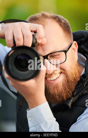 Ein lächelnder Fotograf blickt durch das Kameraobjektiv und hält Momente im Nahaufnahmen-Porträt im Freien fest. Nützlich für Marketing, Werbung, kreative Dienstleistungen, Kommunikation, Fokus, Erfolg, Lifestyle-Konzepte Stockfoto