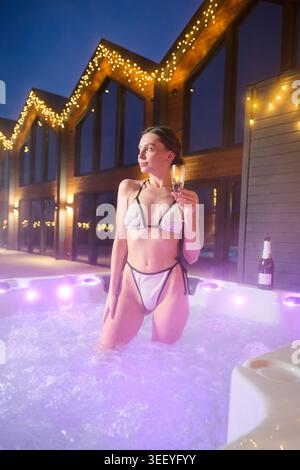 Frau, die sich im dampfenden Whirlpool vor stilvollem Holzhaus, festlicher Beleuchtung und Winterstimmung entspannt, Sekt, Spa Wellness, Komfort und Freizeitkonzept genießen Stockfoto