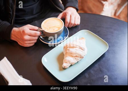 Mannhände halten eine Tasse Kaffee mit Croissant auf Teller im Café, genießen Sie den Moment des Frühstücks, Nahaufnahme von Latte und Gebäck auf dem Tisch, gemütliches Morgenkonzept Stockfoto