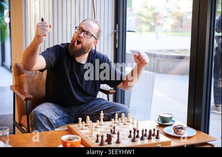 Der Mann feiert das Siegerspiel, während er das Schachstück in einem modernen Wohnzimmer hält. Dramatische Indoor-Szene für Sieg, Erfolg, Strategie, Wettbewerb, Spiel, oder private Freizeitangebote Stockfoto