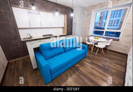 Heller offener Raum mit blauer Couch, Einbauküche und kleinem Essbereich nahe dem Fenster. Moderne Einrichtung des Apartments, intelligentes Layout, Wohnkomfort und Immobilienkonzept Stockfoto