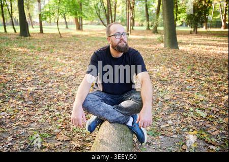 Bärtiger weißer Mann, der auf Baumstamm sitzt, mit nachdenklichem Ausdruck in die Ferne blickt, lässige Haltung zwischen gefallenen Blättern, warmes Nachmittagslicht, besinnliche Lifestyle-Szene für kreative und geistige Gedanken Stockfoto