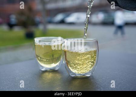 Grüner Tee aus einem Metallkessel auf verschwommenem Hintergrund in ein Glas gießen, heißes Getränk in der Teekanne im japanischen Restaurant, dunkle Tischplatte Stockfoto