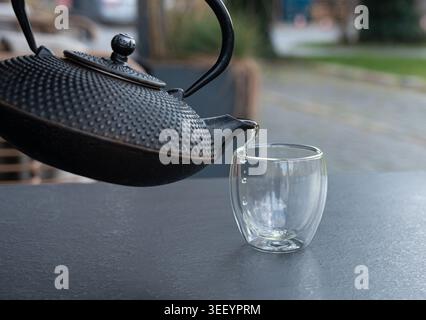 Grüner Tee aus einem Metallkessel auf verschwommenem Hintergrund in ein Glas gießen, heißes Getränk in der Teekanne im japanischen Restaurant, dunkle Tischplatte Stockfoto