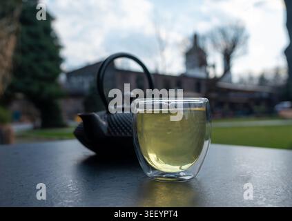 Glasbecher mit grünem Tee und gusseiserner Teekanne auf dunklem Tisch. Traditionelles asiatisches Getränk in modernem doppelwandigen Glas serviert, Teezeit für Menü, ca. Stockfoto