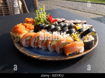 Buntes japanisches Sushi-Set mit verschiedenen Rollen und essbaren Blumen, verschiedene Sushi-Platten mit Lachs, Tempura und frischen Garnelen auf schwarzem Tablet Stockfoto