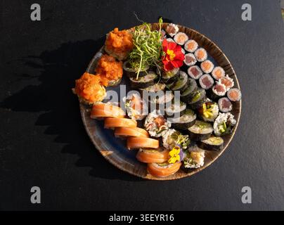 Buntes japanisches Sushi-Set mit verschiedenen Rollen und essbaren Blumen, verschiedene Sushi-Platten mit Lachs, Tempura und frischen Garnelen auf schwarzem Tablet Stockfoto
