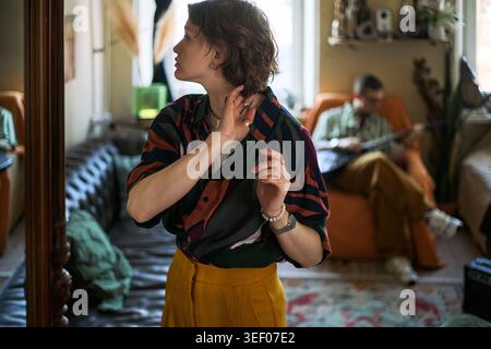 Kaukasische junge Frau, die den Ohrring beim Vorbereiten im Innenraum anpasst, Mann spielt Gitarre im Hintergrund. Repräsentiert alltägliche Lifestyle-Inhalte für Dating, Selbstausdruck, Home Life Marketing Stockfoto