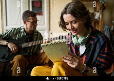 Junge Erwachsene kaukasische Frau schreibt Texte in Notizbuch, während junger Erwachsener kaukasischer Mann Gitarre spielt und zusammen Lied kreiert. Ideal für Musikunterricht, Kreativität und Songwriting Stockfoto