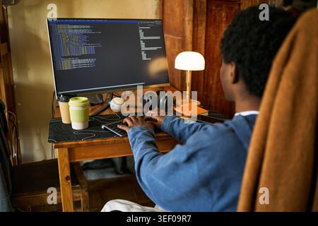 Young Black man codiert am Desktop-Computer im Heimbüro, schreibt Software-Skripte für Fernarbeit und digitale Entwicklung. Ideal für technische Karrieren, Programmier- und freiberufliche Tätigkeiten Stockfoto
