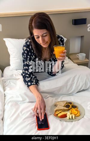 Frau frühstückt im Bett und überprüft ihr Smartphone Stockfoto