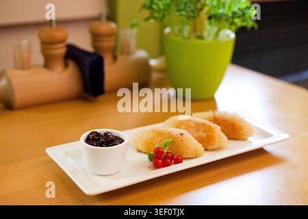 Appetitliches, gebratenes Dessert auf weißem Teller mit Kirschsauce und frischen roten Johannisbeeren. Warmes Licht, geringe Schärfentiefe, gemütliches Restaurant Stockfoto