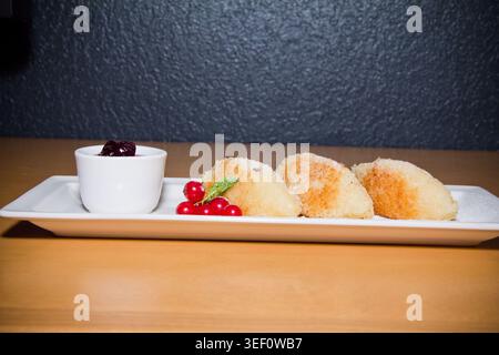 Elegantes Dessert mit goldgebratenem Hüttenkäse, serviert mit süßer Beerensauce und frischen roten Johannisbeeren. Minimalistische Essenspräsentation auf weißem pla Stockfoto