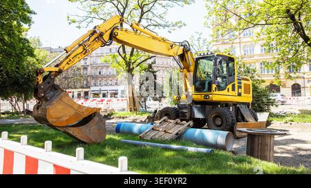 Ein gelber Radbagger, der auf einer Stadtbaustelle mit blauen Wasserleitungen, Sicherheitsbarrieren und städtischen Gebäuden im Hintergrund geparkt ist. Stockfoto