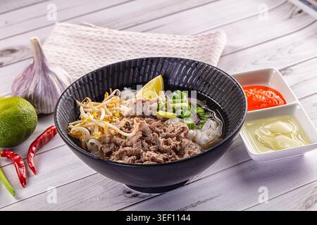 Aromatische Schüssel mit traditionellem vietnamesischem Pho Bo mit Rindfleisch, Nudeln und Sojasprossen. Serviert mit Limette, Chili und Knoblauch auf weißem Holzhintergrund. F Stockfoto