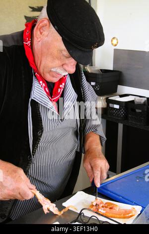 Der alte Mann schneidet vorsichtig Fischfilet. Er trägt ein rotes Bandana und eine schwarze Mütze. Er konzentriert sich auf seine Aufgabe mit leiser Präzision. Dieser Moment fängt eine einfache Sackgasse ein Stockfoto