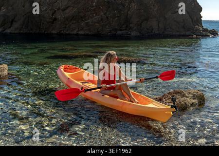 Kajakfahren Frau im Meer Entspannen Sie sich im orangen Kajak auf klarem, ruhigem Meerwasser entlang einer felsigen Küste Stockfoto