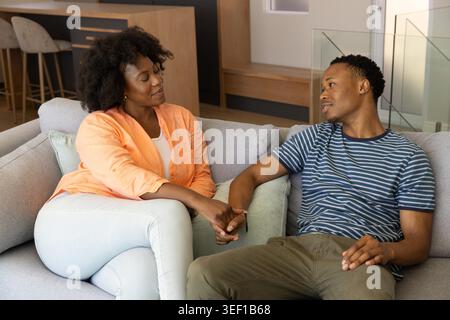 Afroamerikanisches Paar, das im Wohnzimmer auf dem Sofa sitzt und die Hände hält, trägt ein pfirsichfarbenes, gestreiftes T-Stück. Partner, Romantik, zusammen, modern, minimalistisch, warm, neutral Stockfoto