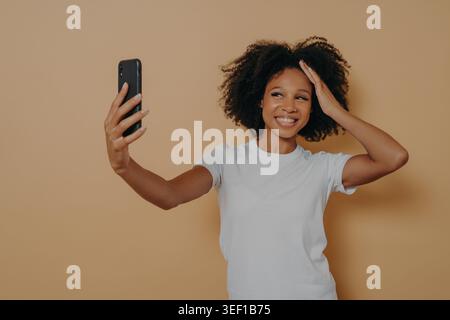 Glückliche charmante dunkelhäutige Frauen 20er in weißem T-Shirt machen Foto-Selfie auf einem modernen Smartphone und lächeln fröhlich, isoliert über beigefarbene Wand. Positive junge afrikanerin, die Fotos auf dem Handy macht Stockfoto