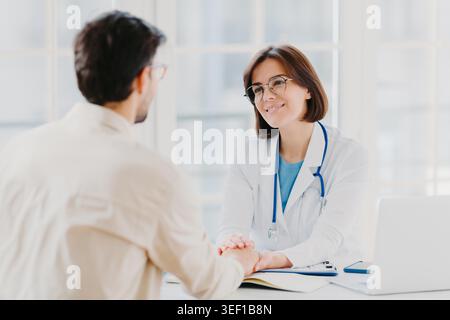Selbstbewusste Ärztin hält Hände von kranken Patienten, überzeugt, dass alles in Ordnung sein wird, in weißem medizinischen Kleid gekleidet, gibt Ratschläge, posiert im Hospialbüro. Beratung und Diagnosekonzept Stockfoto