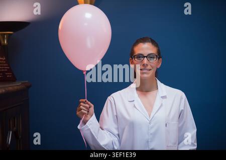 Die Ärztin und Labortechnikerin aus dem Nahen Osten trägt einen weißen Mantel und eine Brille und hält einen rosa Ballon in einem klinischen Bluttestlabor und vermittelt Professionalität, Hoffnung und Mitgefühl. Stockfoto