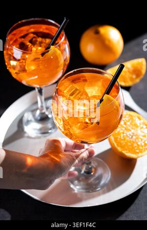 Nahaufnahme einer Frauenhand mit einem Aperol Spritz Cocktail serviert mit Eiswürfeln, Orangenscheiben und schwarzem Strohhalm. Stockfoto