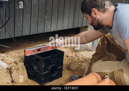 Ein Mann installiert ein Regenwasser-Auffangbecken für das Entwässerungssystem eines Landhauses und überprüft seine Position mit einer Wasserwaage. Hochwertige Fotos Stockfoto
