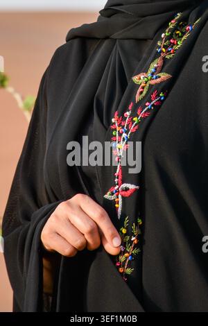 Detaillierte Nahaufnahme einer Frauenhand auf einer schwarzen Abaya mit farbenfroher Blumenstickerei und Perlen in einer Wüstenumgebung. Stockfoto
