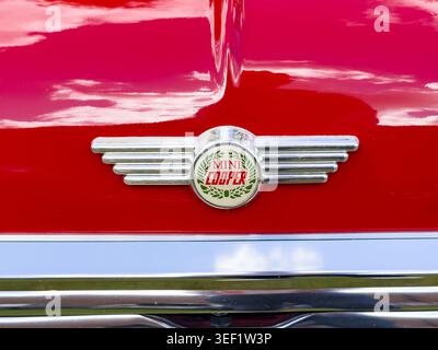 Nahaufnahme eines roten Minicooper-Autoemblems mit verchromten Flügeln auf einer glänzend roten Motorhaube Stockfoto